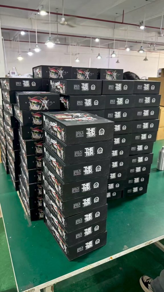 Stacks of black boxes labeled 'THE TEN' stacked on a green production table in a warehouse.