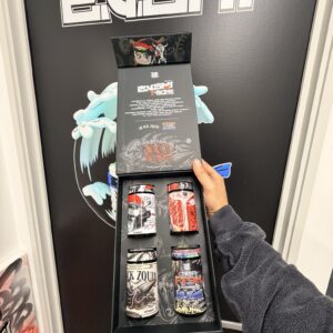 Person holds a black display stand with illustrated cans featuring bold red, black, and white artwork in front of a dark wall with white graphic text behind.