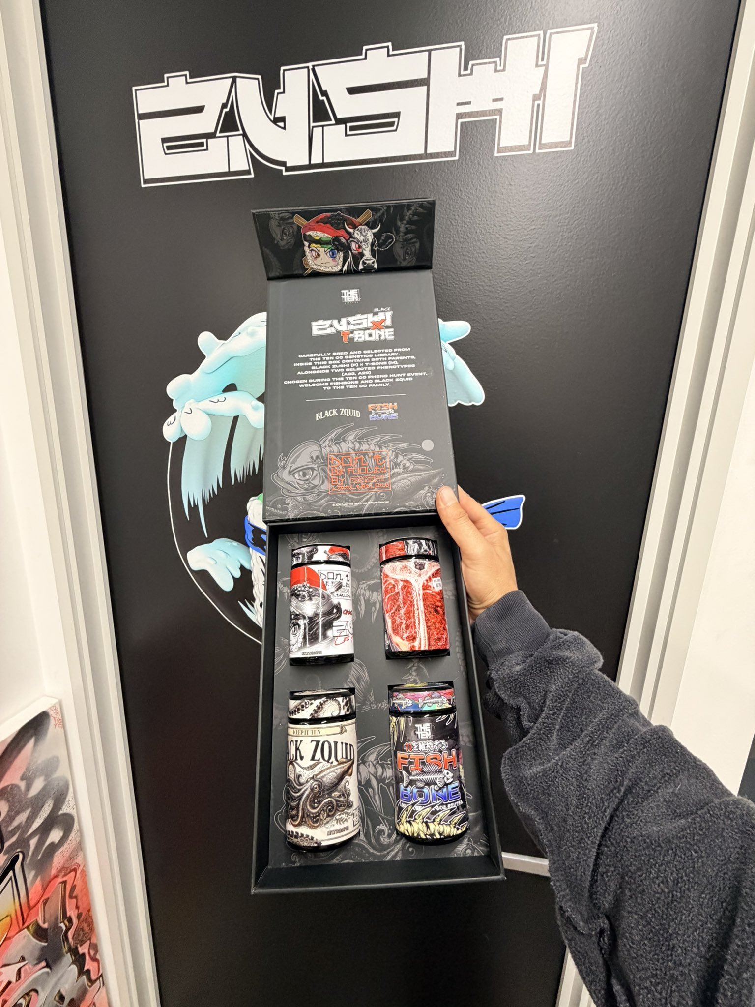 Person holds a black display stand with illustrated cans featuring bold red, black, and white artwork in front of a dark wall with white graphic text behind.