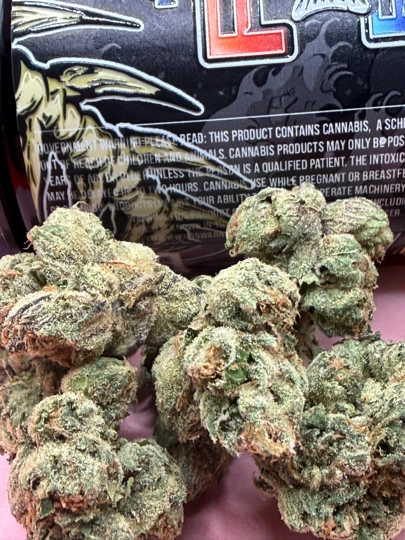 Close-up of frosty cannabis buds with green and purple hues in front of a dark, text-labeled package image behind them.